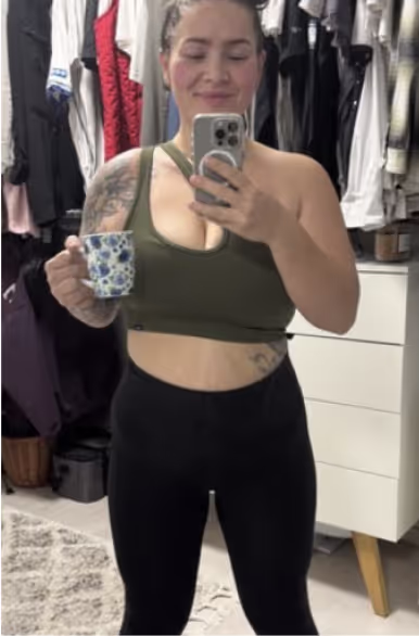 Eine lächelnde Frau in Sportkleidung macht ein Selfie im Spiegel und hält eine Tasse.