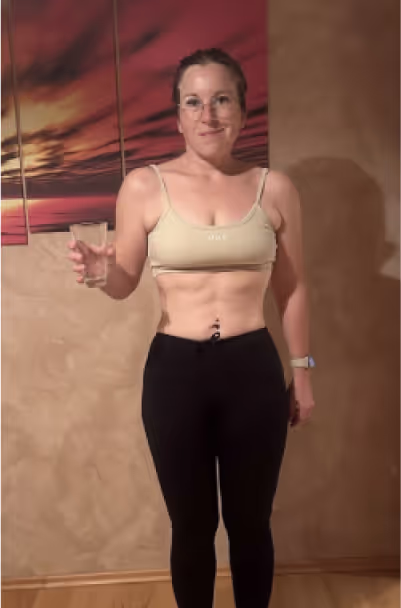 Eine lächelnde Frau in Sportkleidung hält ein Glas Wasser in der Hand.