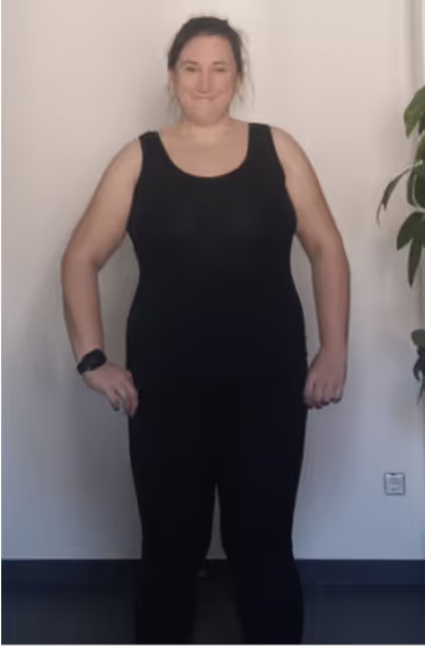 Eine lächelnde Frau in schwarzem Tanktop und Leggings steht vor einer weißen Wand.