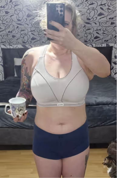 Eine Frau in Sport-BH und Shorts macht ein Selfie im Spiegel und hält eine Tasse.