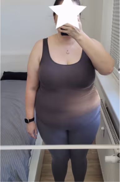 Eine Frau in einem Tanktop und Leggings macht ein Selfie im Spiegel, ihr Gesicht ist mit einem Stern-Emoji bedeckt.